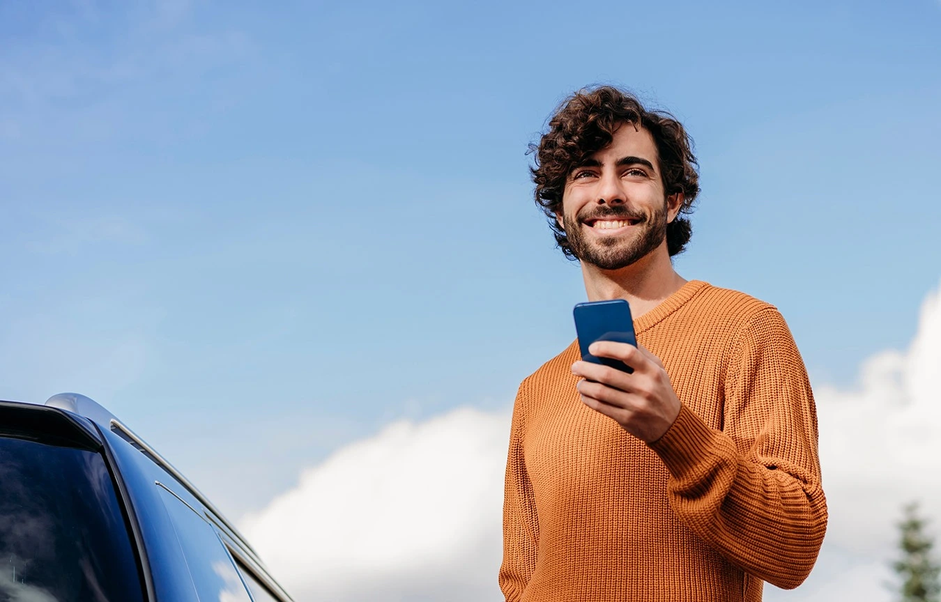 Giovane uomo con smartphone in mano accanto automobile Young man holding smartphone beside car